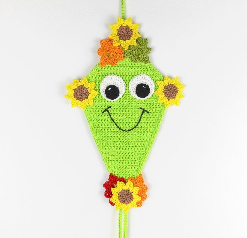 Lime green crocheted kite-shaped hanging decoration with big eyes, stitched smile and yellow-orange flower appliqués