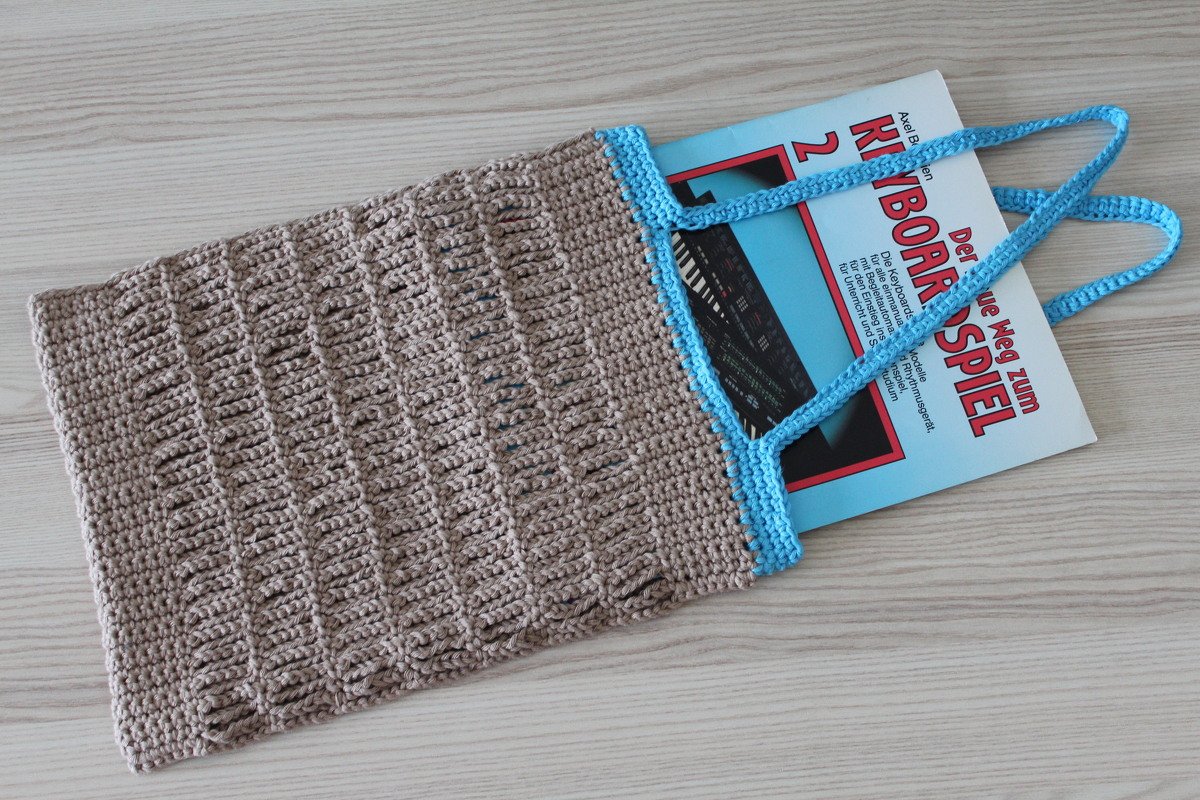 Beigefarbene gehäkelte Tasche mit hellblauem Rand und Henkeln auf Holz, mit einem Buch teilweise darin.