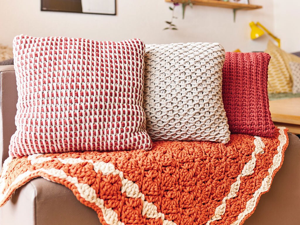 Nahaufnahme von drei gehäkelten Kissen in Rot, Weiß und Beige sowie einer orangefarbenen Häkeldecke auf einem Sofa.