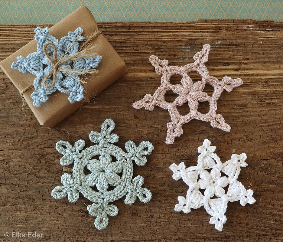 Vier gehäkelte Schneeflocken in pastellfarben und weiß auf Holz, eine davon auf einem Geschenk