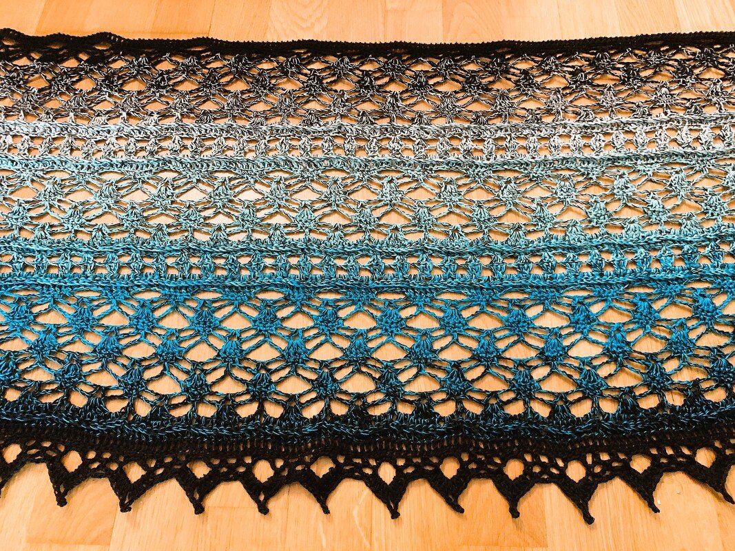 Ombre crocheted scarf in black, grey and blue with open lace stitch on wooden floor