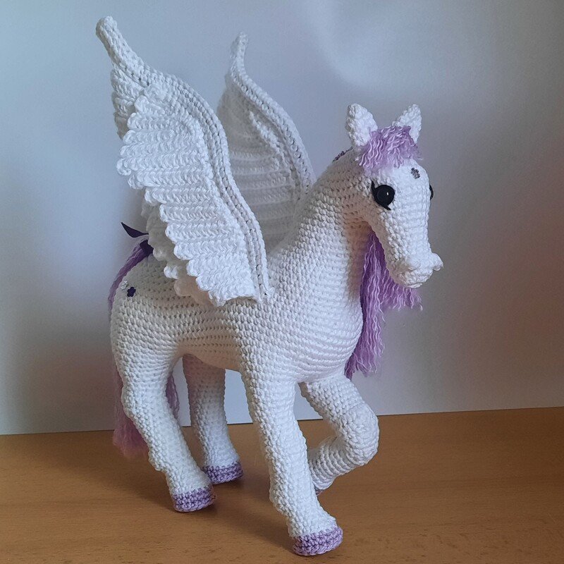 Weißer gehäkelter Pegasus mit großen Flügeln und lilafarbener Mähne steht auf einer Holzoberfläche vor hellem Hintergrund.