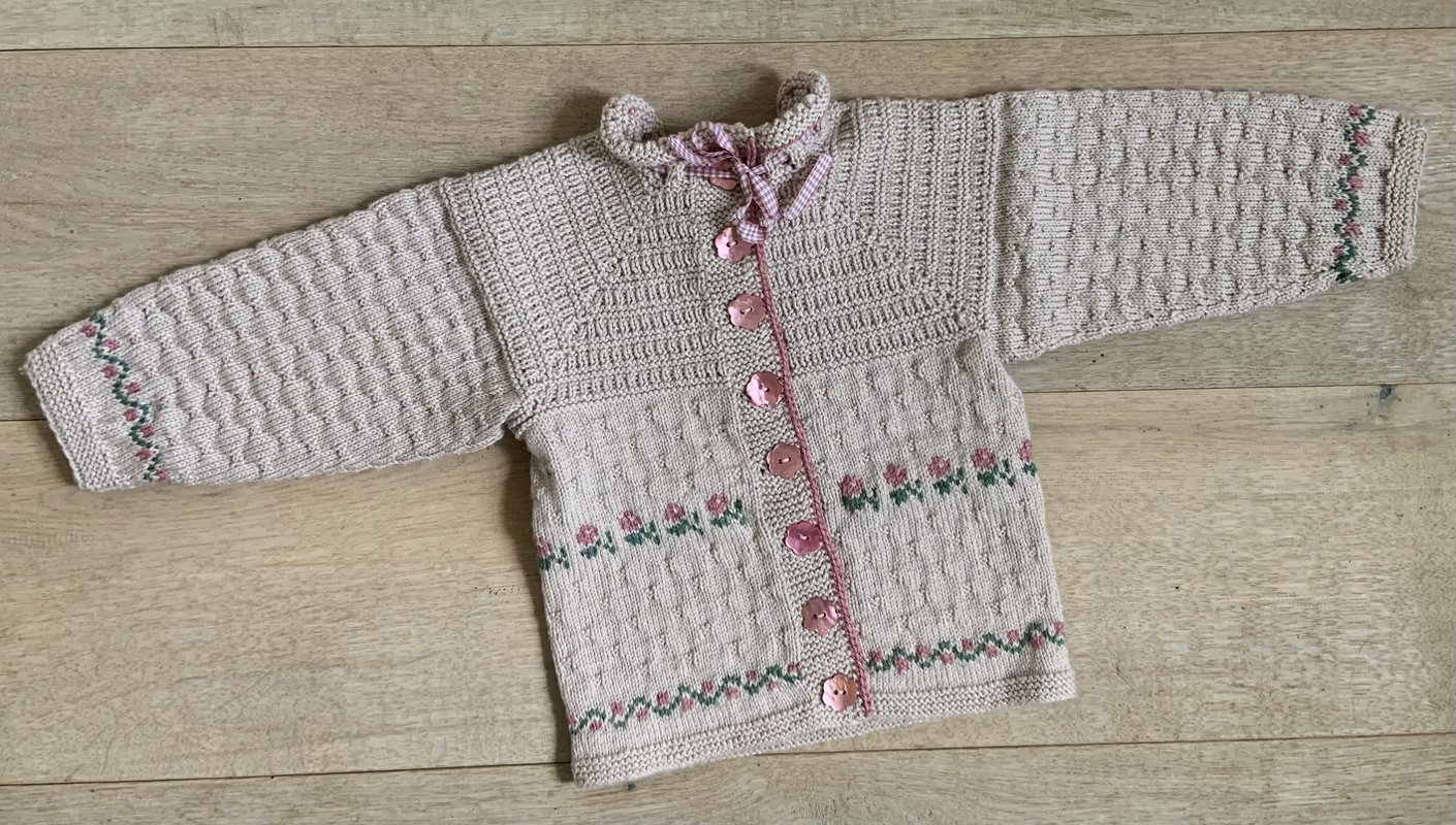 Rosa gestrickte Babyjacke mit Blumenbordüre und rosafarbenen blütenförmigen Knöpfen, flach auf Holzuntergrund gelegt