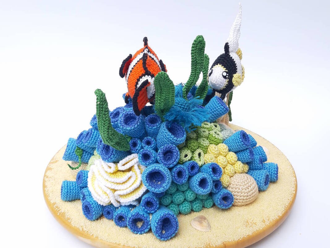Amigurumi coral reef diorama with crocheted clownfish, bannerfish, and colorful yarn corals on a sandy base.
