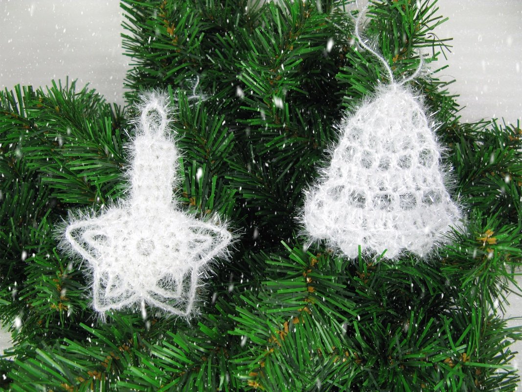 Zwei weiße gehäkelte Weihnachtshänger (Stern mit Kerzenmotiv und Glocke) auf grünen Tannenzweigen.