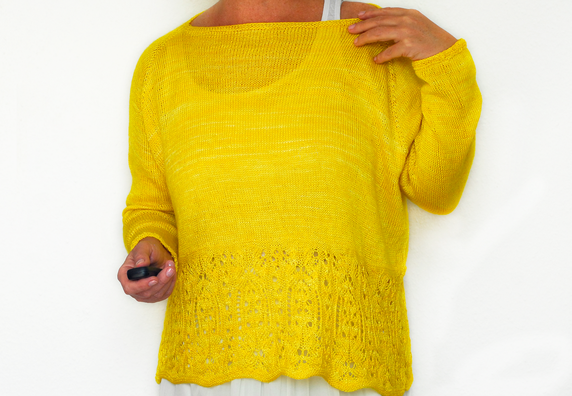 Nahaufnahme eines gelben gestrickten Pullovers mit Lochmuster am unteren Saum und weitem U-Boot-Ausschnitt. Eine Hand hält ein kleines schwarzes Objekt.