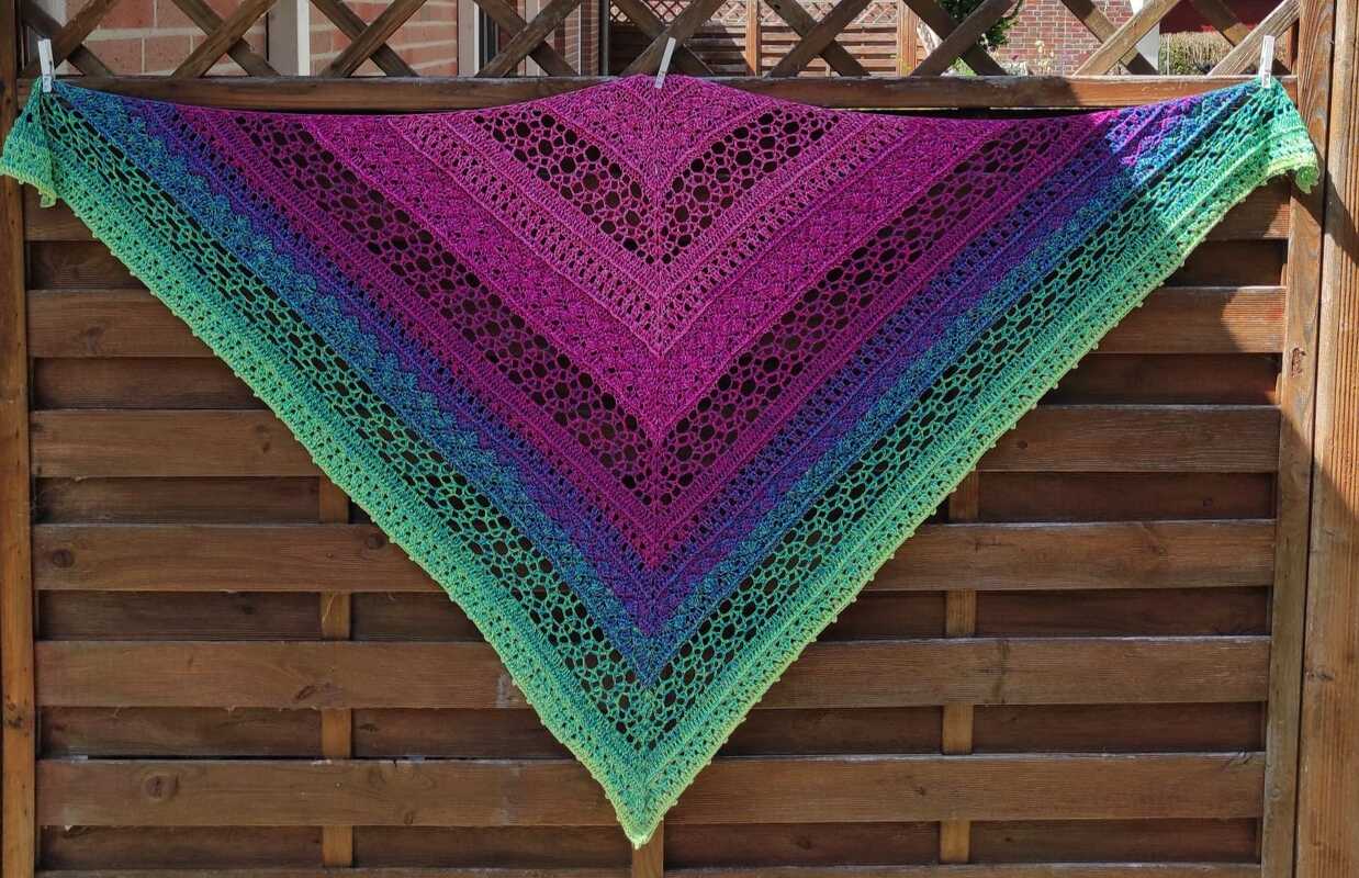 Crocheted triangular shawl with an ombre gradient from green to blue, purple, and magenta, featuring openwork stitches, hung on a wooden fence.