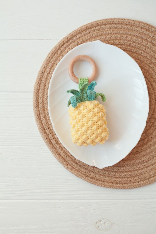 Crocheted yellow pineapple baby teether with green leaves on a wooden ring, placed on a white plate over a woven mat