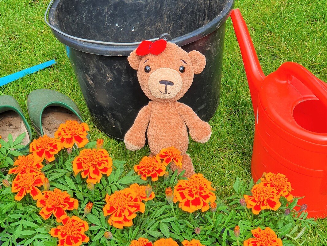 Brauner Amigurumi-Teddybär mit roter Schleife im Gras zwischen orangefarbenen Blumen, neben roter Gießkanne und schwarzem Eimer.