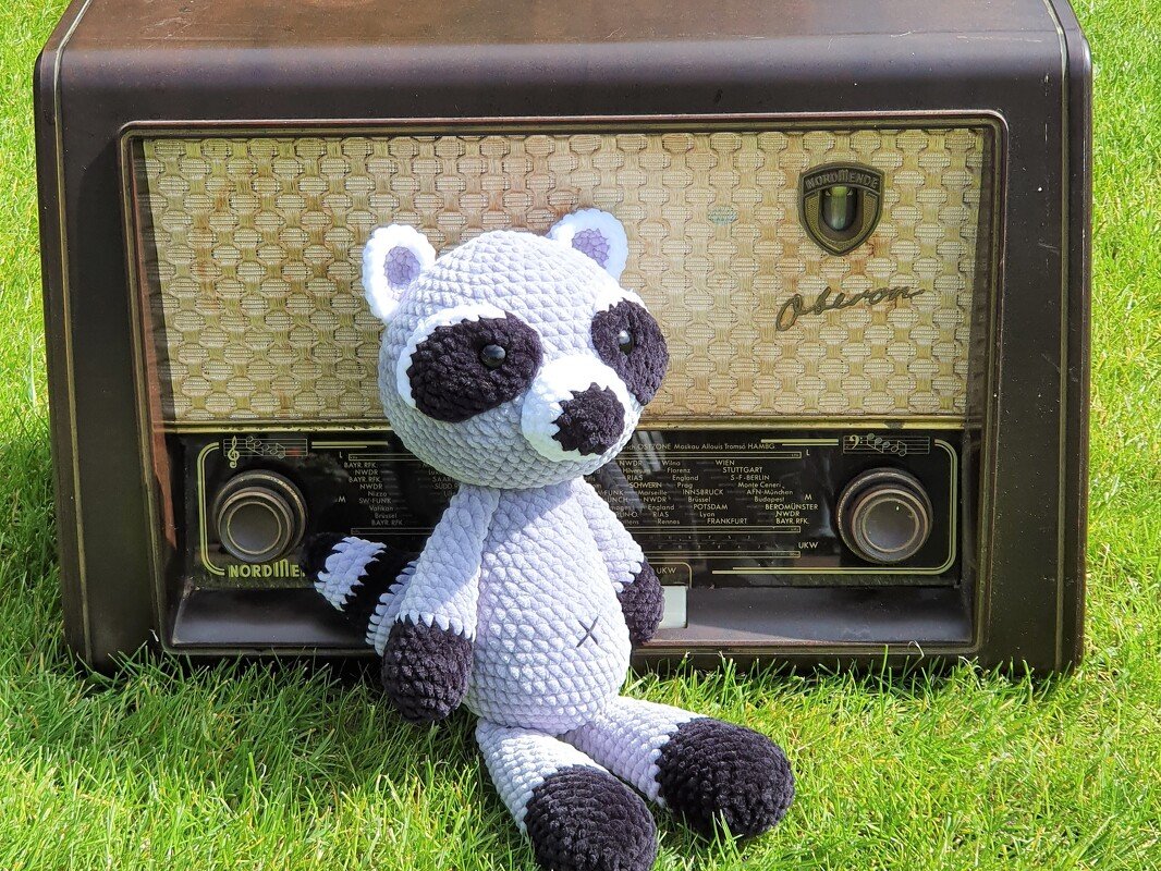 Gehäkelter Amigurumi-Waschbär in Grau und Schwarz sitzt auf Gras vor einem alten Radio.