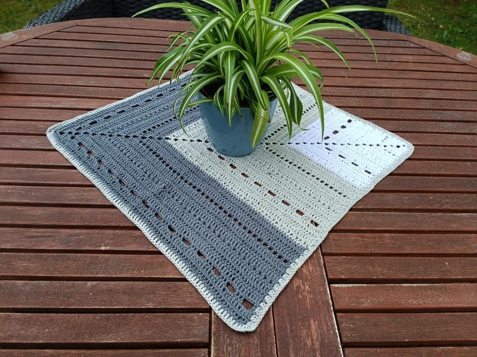 Quadratisches gehäkeltes Deckchen in Dunkelgrau, Hellgrau und Weiß auf einem Holztisch unter einem Blumentopf