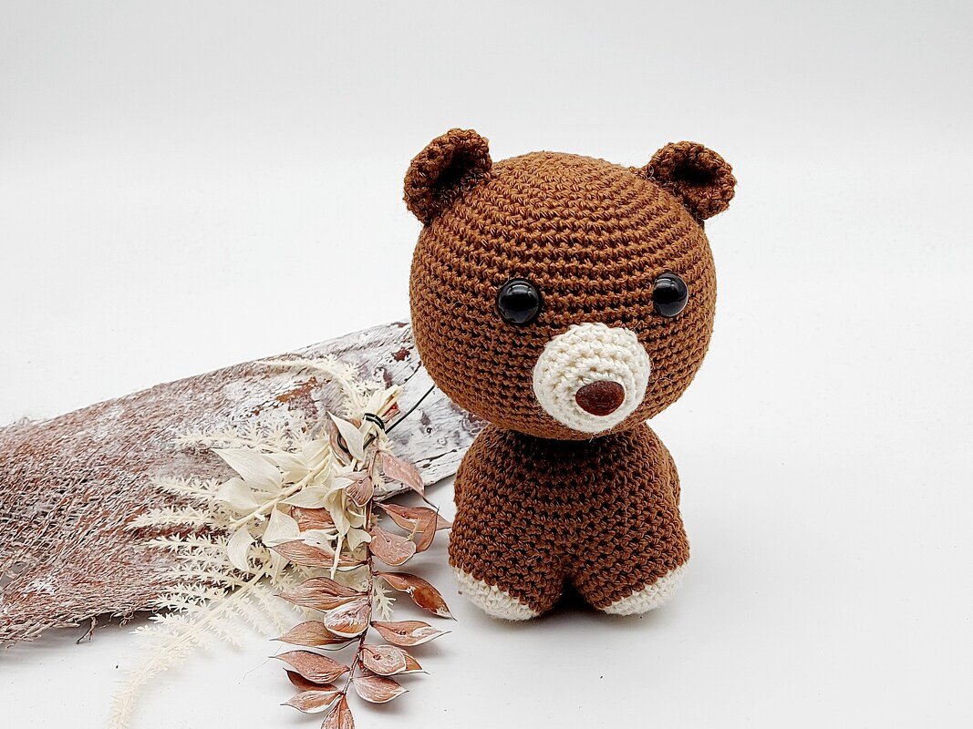 Gehäkelter brauner Teddy als Amigurumi neben Trockenblumen und Rinde auf hellem Hintergrund