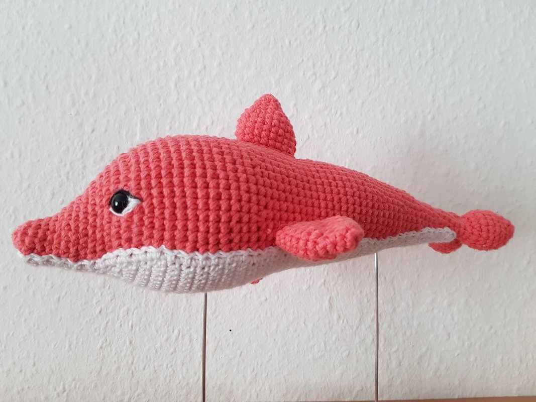 Korallenrotes gehäkeltes Delfin-Amigurumi mit weißem Bauch und schwarzem Auge vor weißer Wand