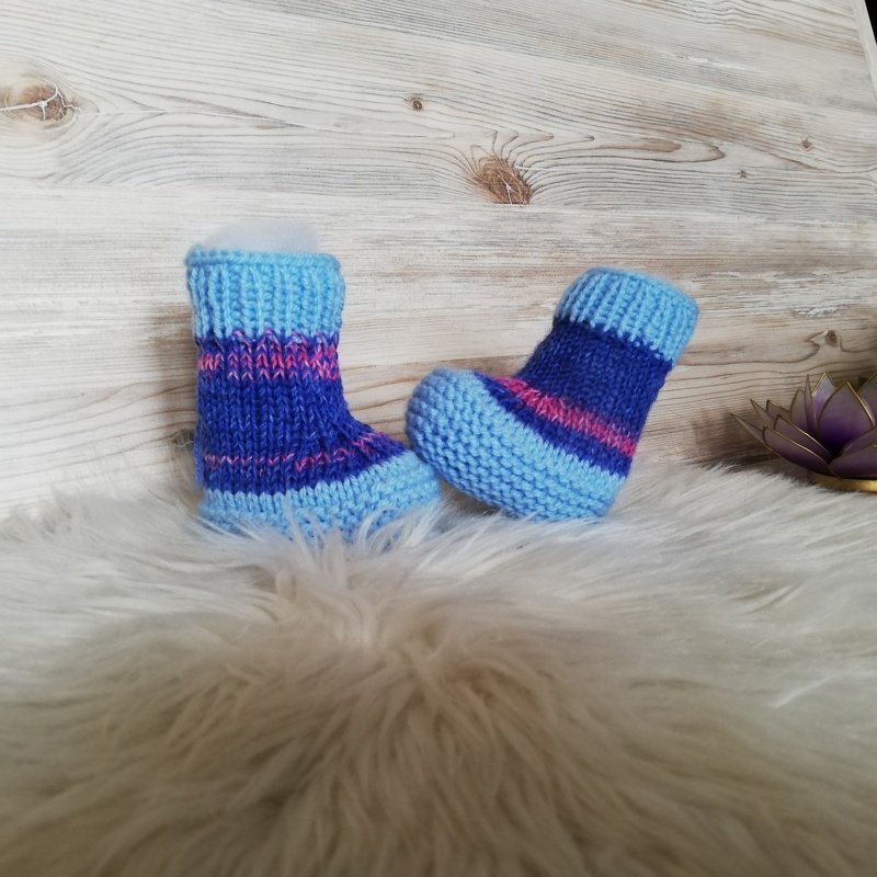 Zwei gestrickte Babysocken in Hellblau, Dunkelblau und Pink auf weißem, flauschigem Untergrund vor einer Holzleiste.