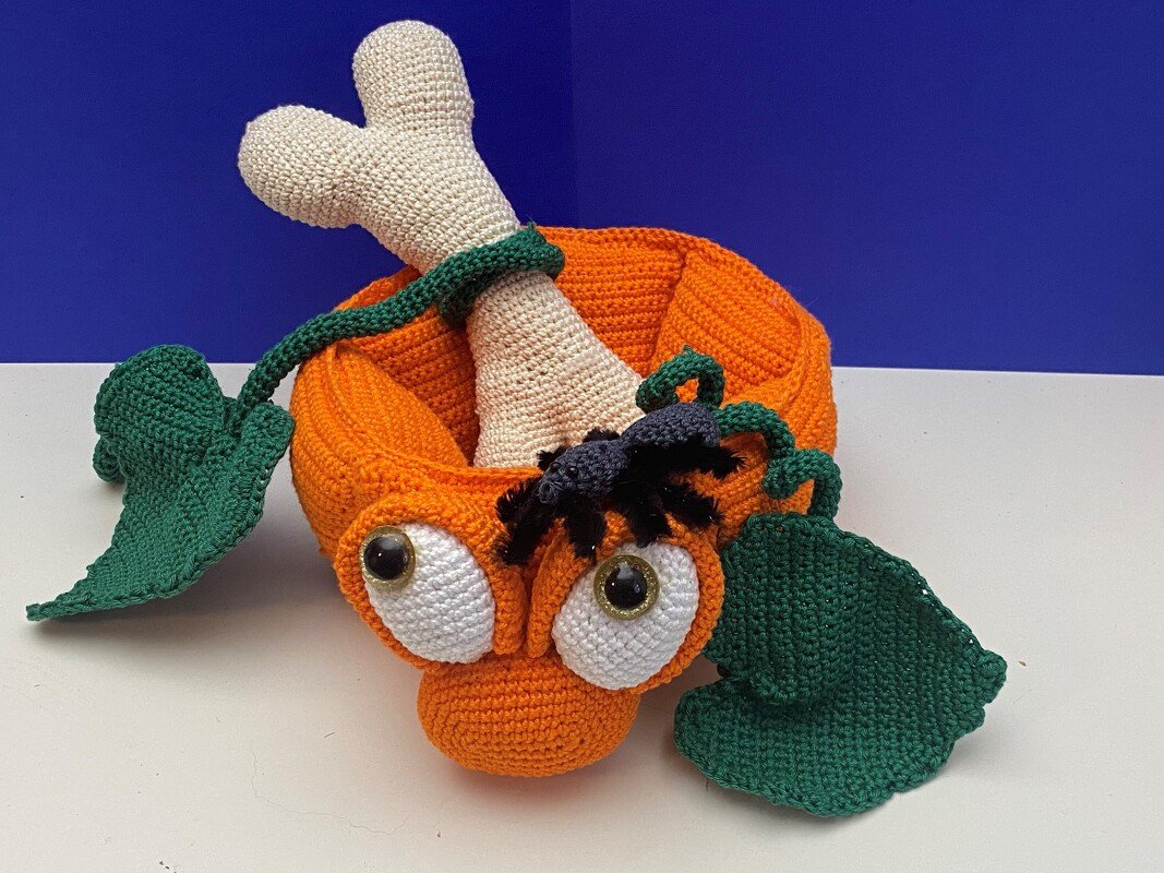 Gehäkelte Halloween-Kürbis-Schale mit großen Augen, grünen Blättern, kleiner Spinne und einem Knochen, der aus der Öffnung ragt