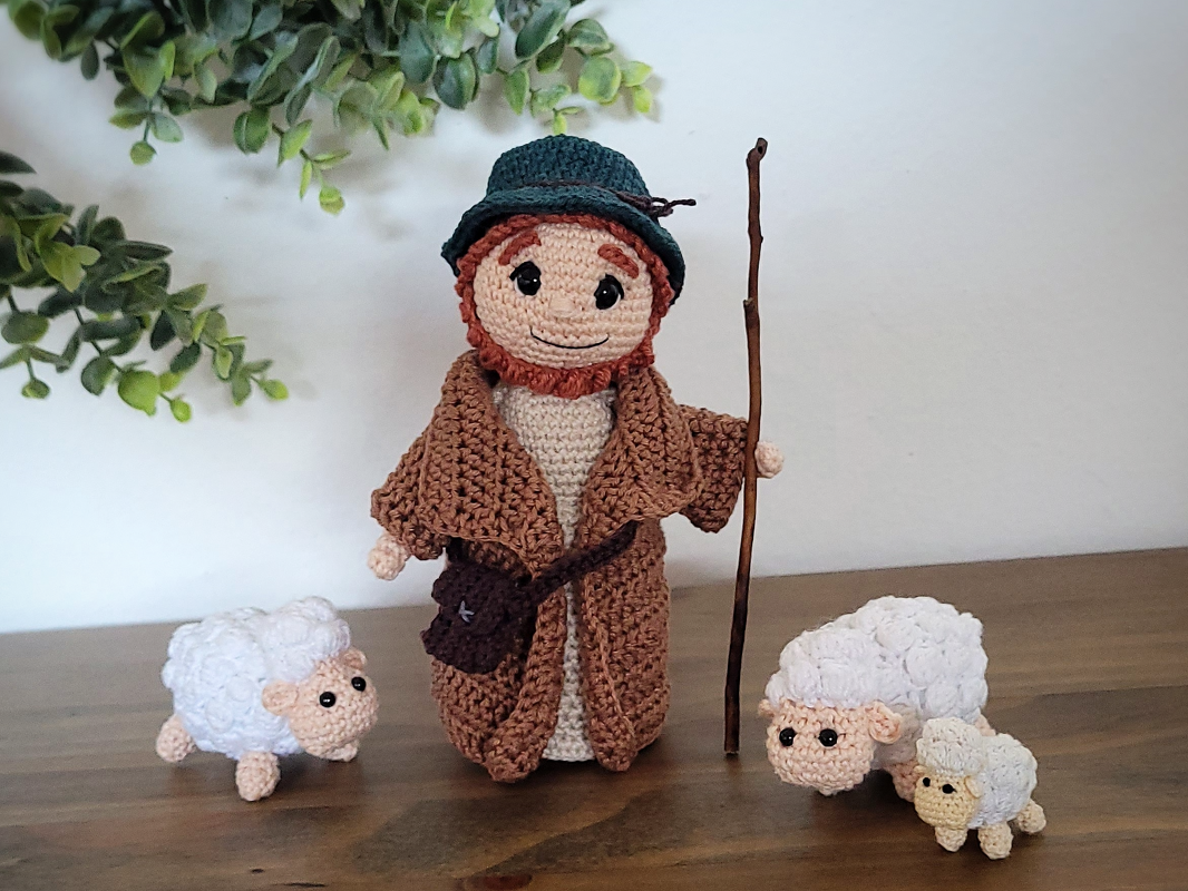 Gehäkelte Amigurumi-Puppe als Hirte mit Hut und Stab, daneben drei kleine gehäkelte Schafe auf einem Holztisch