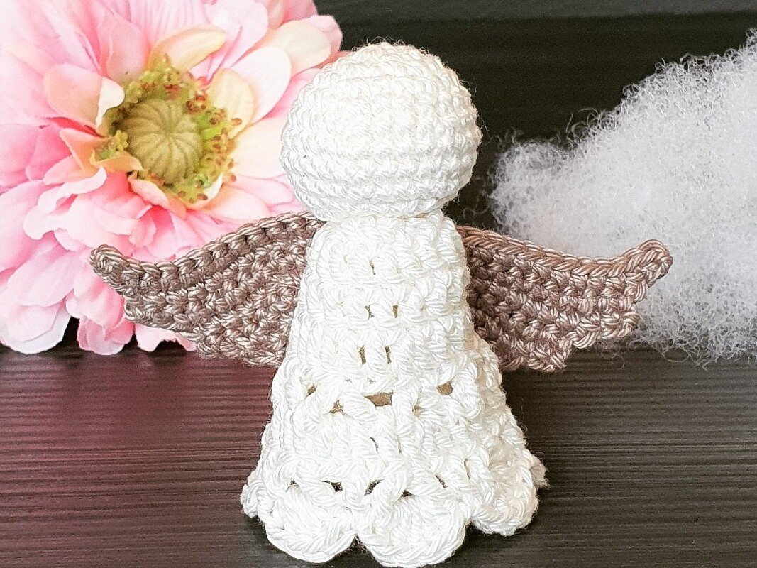 Rückansicht eines weißen Amigurumi-Engelchens mit braunen Flügeln neben einer rosa Blüte und Watte