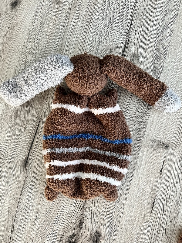 Gestricktes braunes Schmusetuch mit weißen, grauen und blauen Streifen auf hellem Holz.