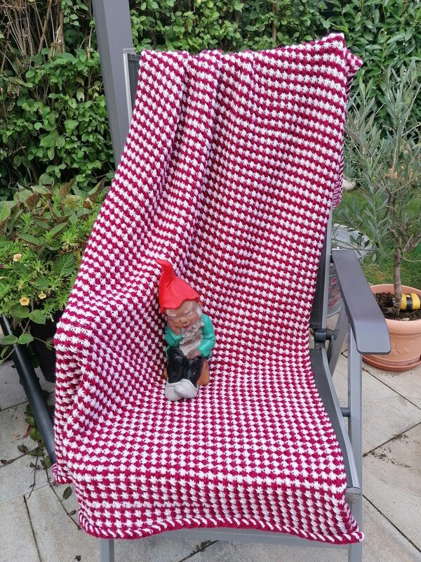 Gestrickte rot-weiße Decke mit Hahnentritt-Muster auf Gartenstuhl mit Gartenzwerg