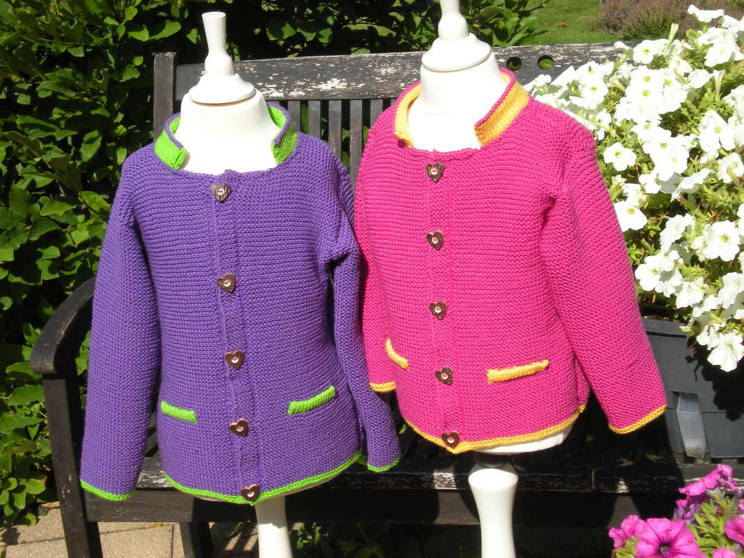 Two knitted children's cardigans on dress forms: purple with green trim and pink with yellow trim, shown outdoors on a bench.