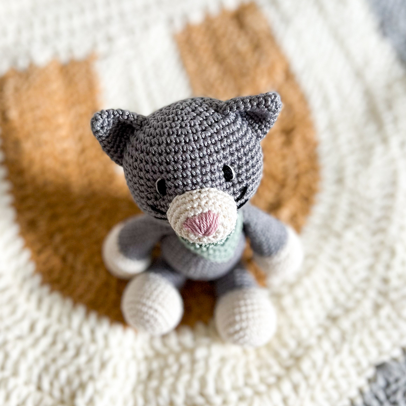 Grau-weißes gehäkeltes Amigurumi-Kätzchen mit hellgrünem Schal auf weiß-brauner Häkeldecke