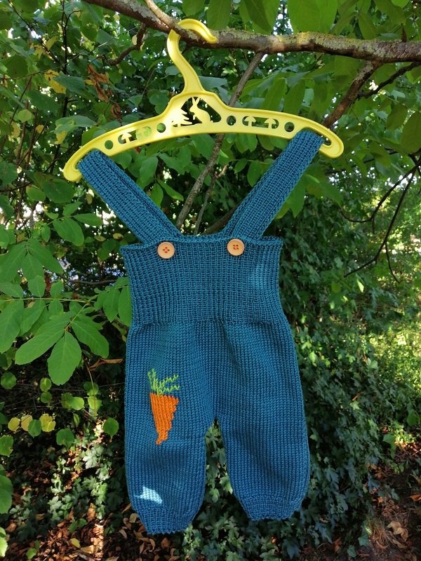 Petrolfarbene gestrickte Baby-Latzhose mit Karotten-Applikation und Holzknöpfen an gelbem Kleiderbügel