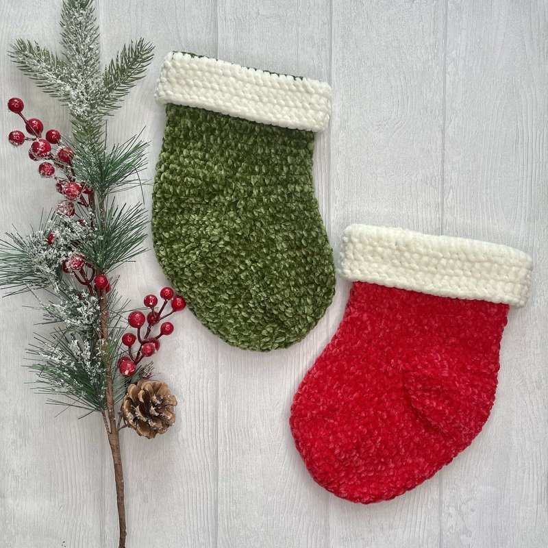 Zwei gehäkelte Weihnachtsstrümpfe, einer rot und einer grün, mit weißem Umschlag neben Tannenzweig, roten Beeren und Zapfen auf hellem Holz