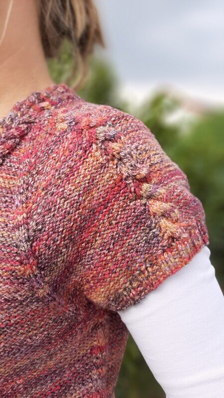 Nahaufnahme eines mehrfarbigen Kurzarm-Strickpullovers mit Zopfmustern an Schulter und Ärmel, kombiniert mit weißem Langarmshirt.