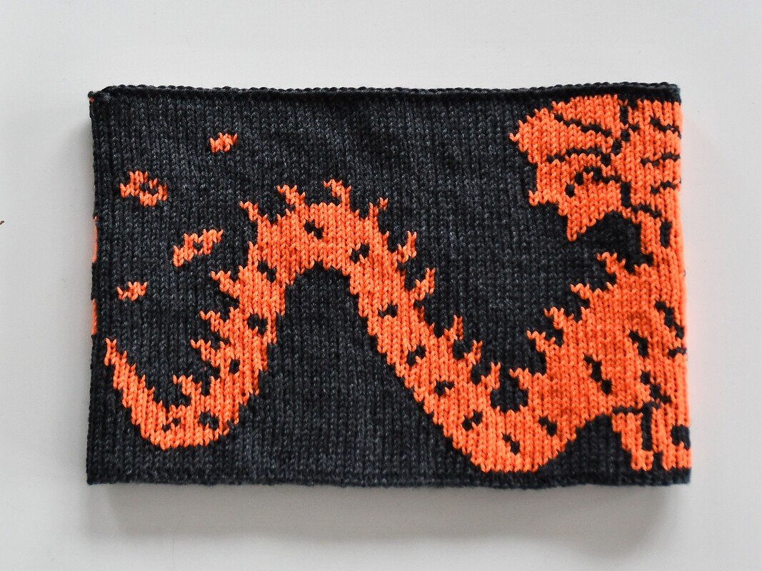 Rectangular knitted scarf or cowl with a bold orange flame-like pattern on a dark gray background.
