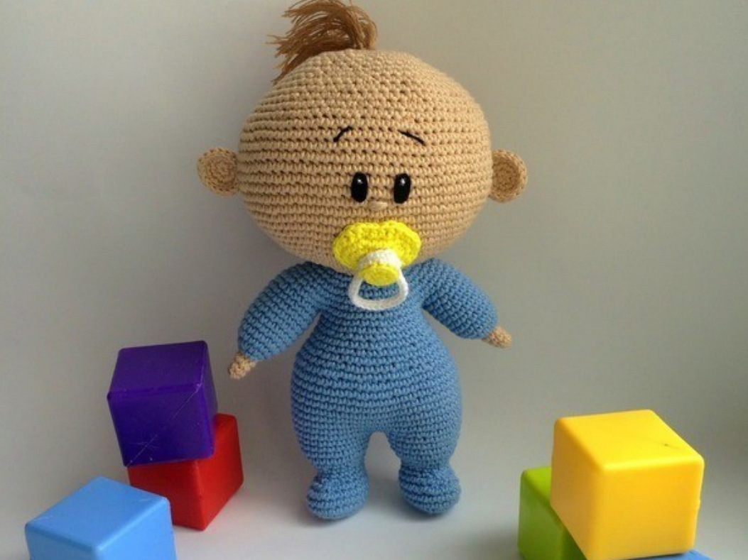 Amigurumi-Babypuppe mit hellem Gesicht, blauem Strampler und gelbem Schnuller, umgeben von bunten Bauklötzen.
