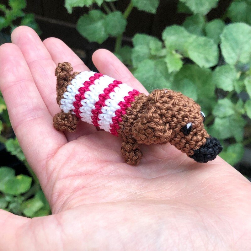 Kleiner gehäkelter Dackel in Braun mit rot-weiß gestreiftem Pulli, liegend in einer Hand vor grünem Hintergrund