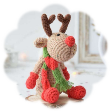 Sitzendes gehäkeltes Rentier-Amigurumi mit roter Nase, grünem Schal mit Herzmotiven und roten Handschuhen vor hellem Hintergrund.