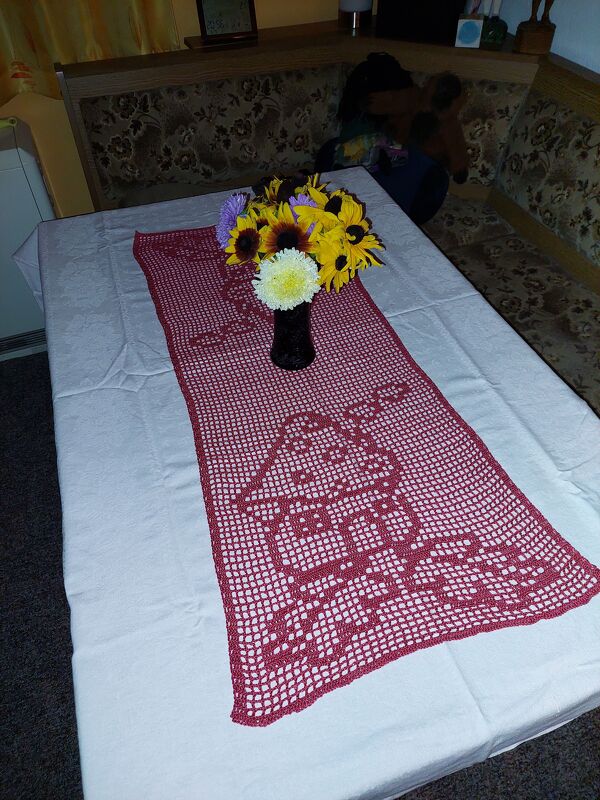 Roter gehäkelter Tischläufer mit Hausmotiv auf weißer Tischdecke und Vase mit Sonnenblumen