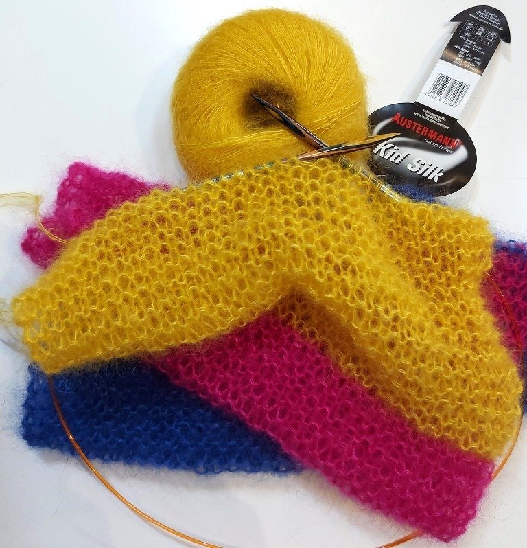 Gelbes flauschiges Garnknäuel, Rundstricknadel und mehrfarbiges gestricktes Stück in Gelb, Pink und Blau auf hellem Hintergrund.