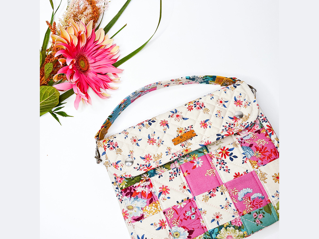 Gesteppte Utensilo-Tasche im Patchwork-Stil mit floralen Mustern in Weiß, Rosa und Blau, neben einer einzelnen pinken Blüte