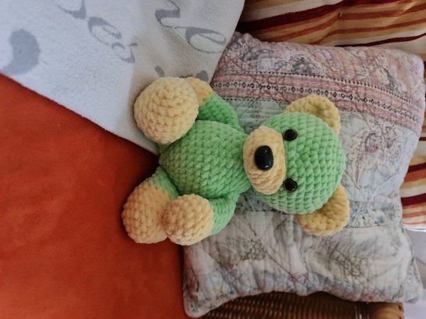 Gehäkelter Amigurumi-Teddybär in Hellgrün mit gelben Ohren und Pfoten, liegend auf einem Sofa.