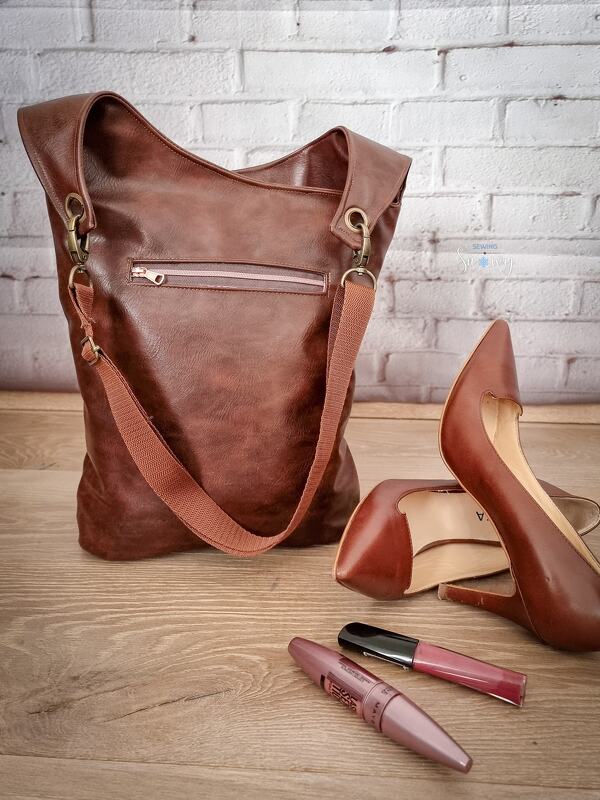 Braune Tasche mit außenliegendem Reißverschluss, Metallbeschlägen und Schulterriemen neben braunen Pumps und Make-up auf Holzboden.