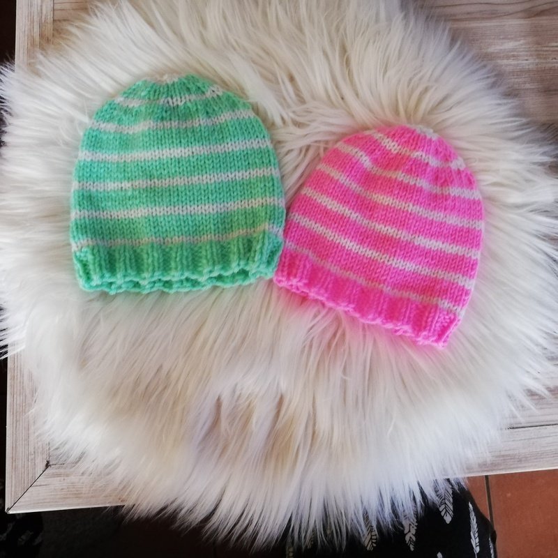 Zwei gestrickte Babymützen, mint-weiß und pink-weiß gestreift, mit geripptem Bund auf hellem Fell