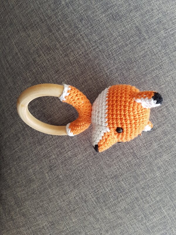 Gehäkelter orange‑weißer Fuchskopf an rundem Holzring auf grauem Stoff