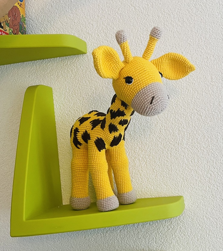 Gehäkelte gelbe Amigurumi-Giraffe mit dunklen Flecken auf grünem Wandregal