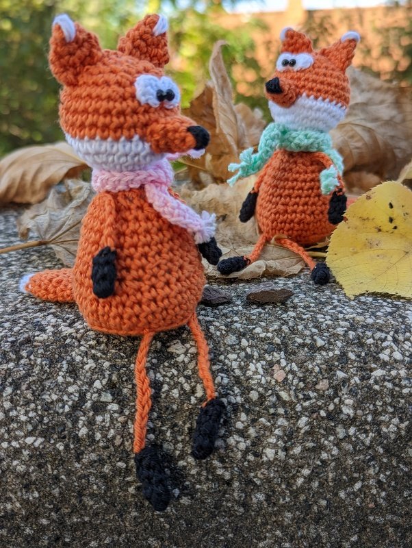 Zwei orange-weiße gehäkelte Fuchs-Amigurumi mit Schals sitzen auf einem Stein vor Herbstblättern