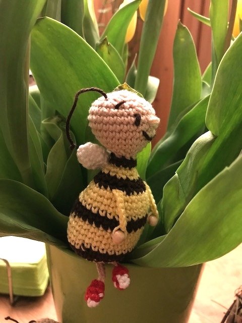 Gehäkelte Amigurumi-Biene mit gelb-schwarzen Streifen und roten Schuhen im grünen Pflanztopf.