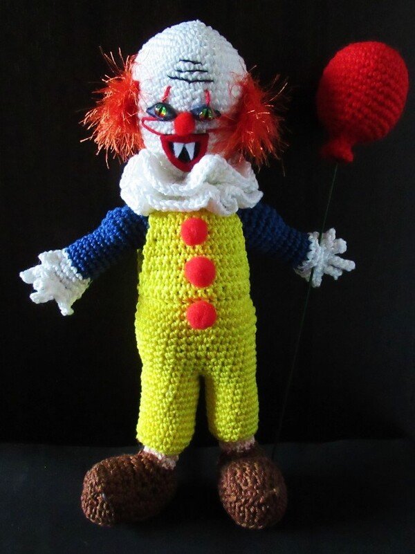 Gehäkelte Clownpuppe mit weißem Kopf, orangefarbenem Haar, gelber Latzhose und rotem Ballon vor schwarzem Hintergrund