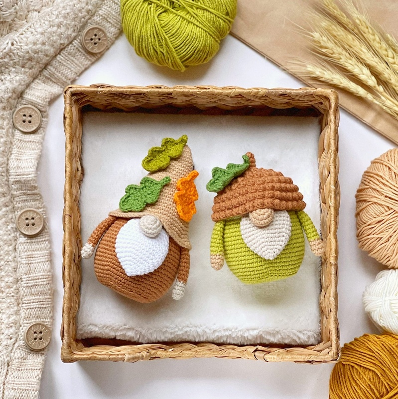 Two crocheted amigurumi autumn gnome dolls with leaf-decorated hats and white beards in a wicker tray, surrounded by yarn