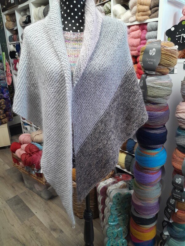 Gestricktes Schultertuch mit Grau-Farbverlauf auf einer Schaufensterpuppe, Garnregale im Hintergrund.