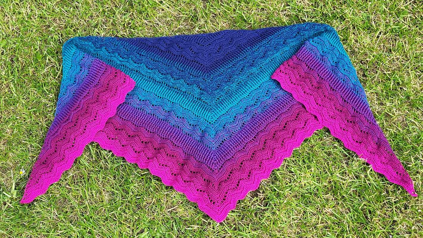 Gehäkeltes Dreieckstuch in Blau, Türkis, Lila und Pink mit Wellenmuster auf Gras.
