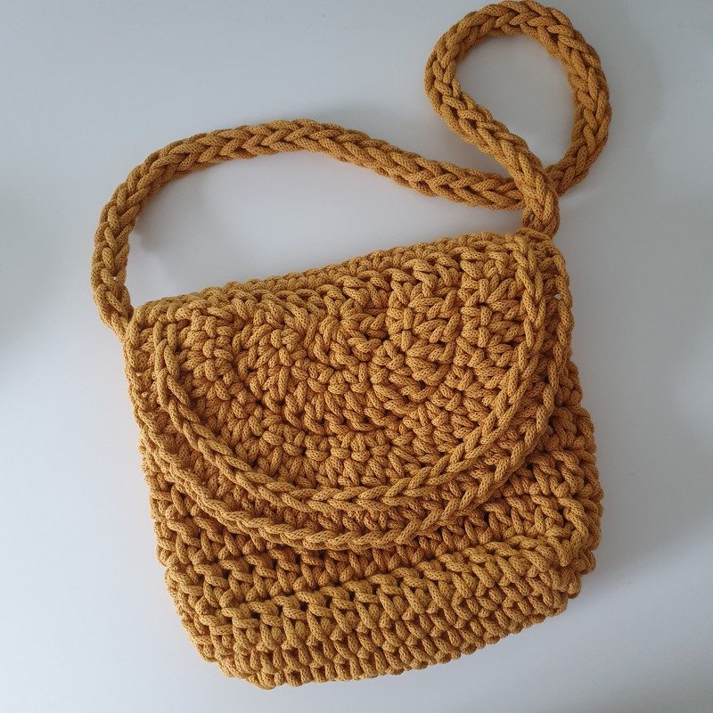 Gehäkelte senfgelbe Umhängetasche mit Klappenverschluss und geflochtenem Gurt auf weißem Hintergrund