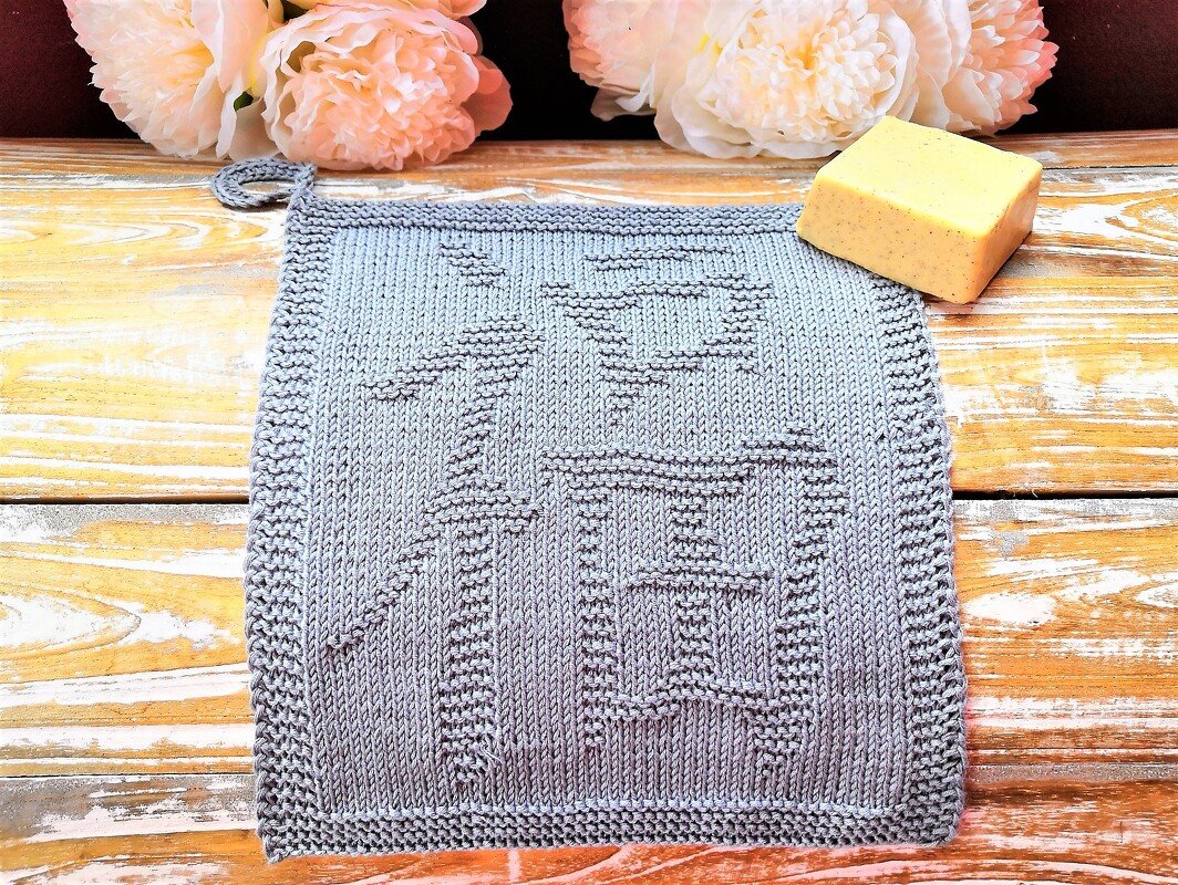 Light blue-gray knitted potholder with raised character motif and corner loop on wooden surface with flowers and soap.