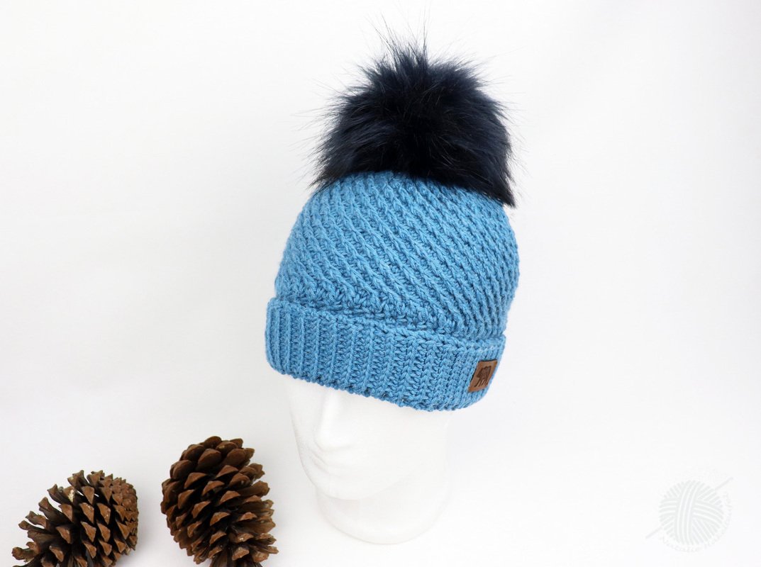 Hellblaue gehäkelte Mütze mit dunklem Bommel und Rippenmuster auf weißem Kopfmodell, Tannenzapfen im Vordergrund