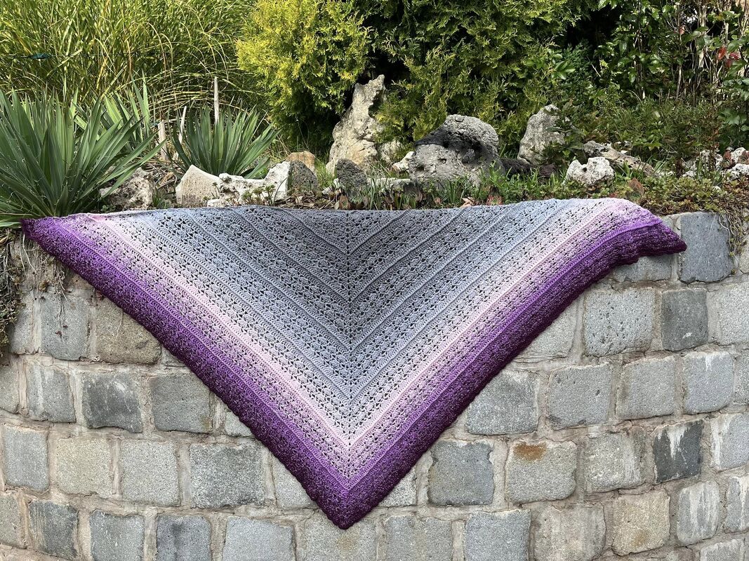 Gehäkeltes Dreieckstuch in Grau, Rosa und Lila mit Farbverlauf und Spitzenmuster auf einer Steinmauer